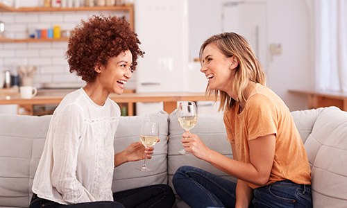 Two women sitting on the couch lauhging and drinking wine.